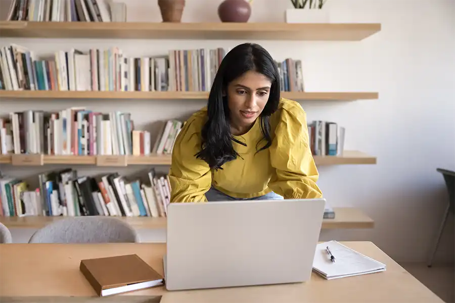 woman standing at workplace table, leaning over laptop typing
