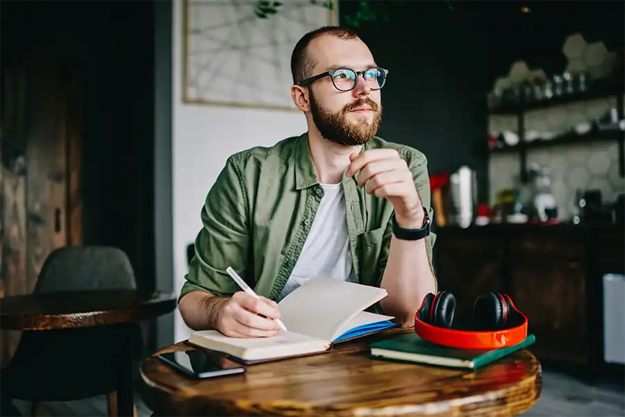 A red headed middle aged man with glasses wearing a green button up is sitting in a cafe at a small wooden table. He is looking off into the distance while holding a white pen hovering over an open notebook.